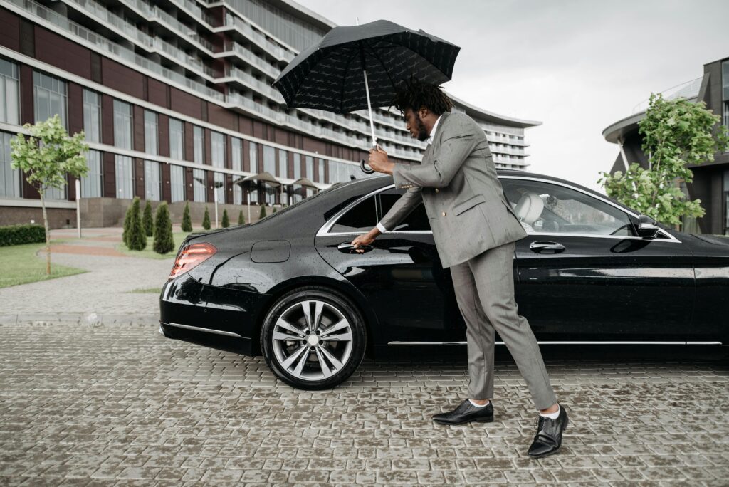 Elegant man in suit opens car door under umbrella by modern building on rainy day.